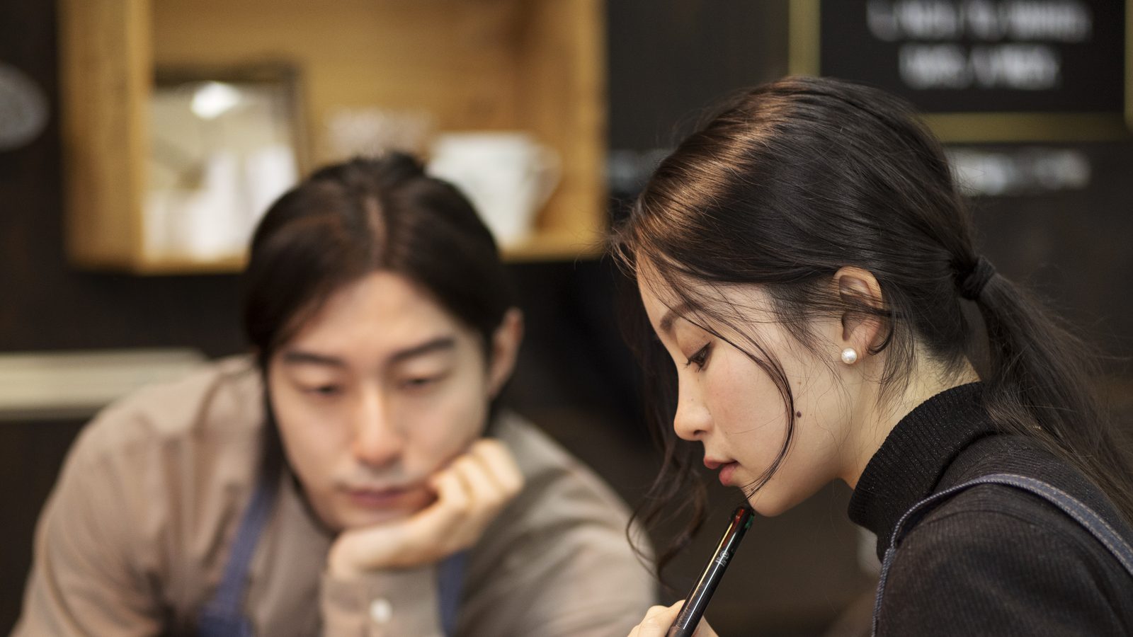 Two café staff at the counter, one reading notes while the other works at a laptop